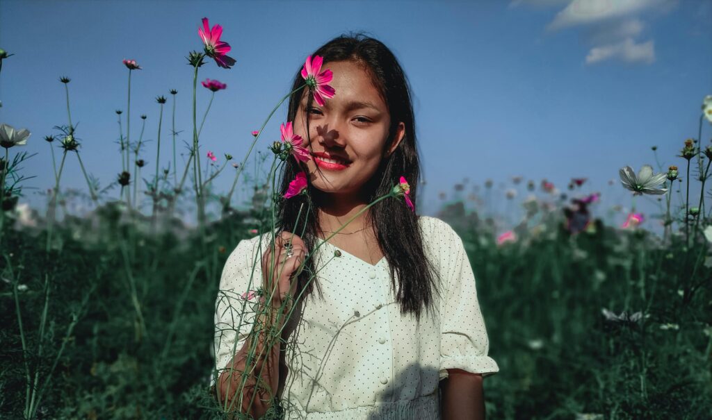 Positive young ethnic female in white dress holding stems of flowers putting buds to face while standing among blooming plantation against blue sky in sunny day